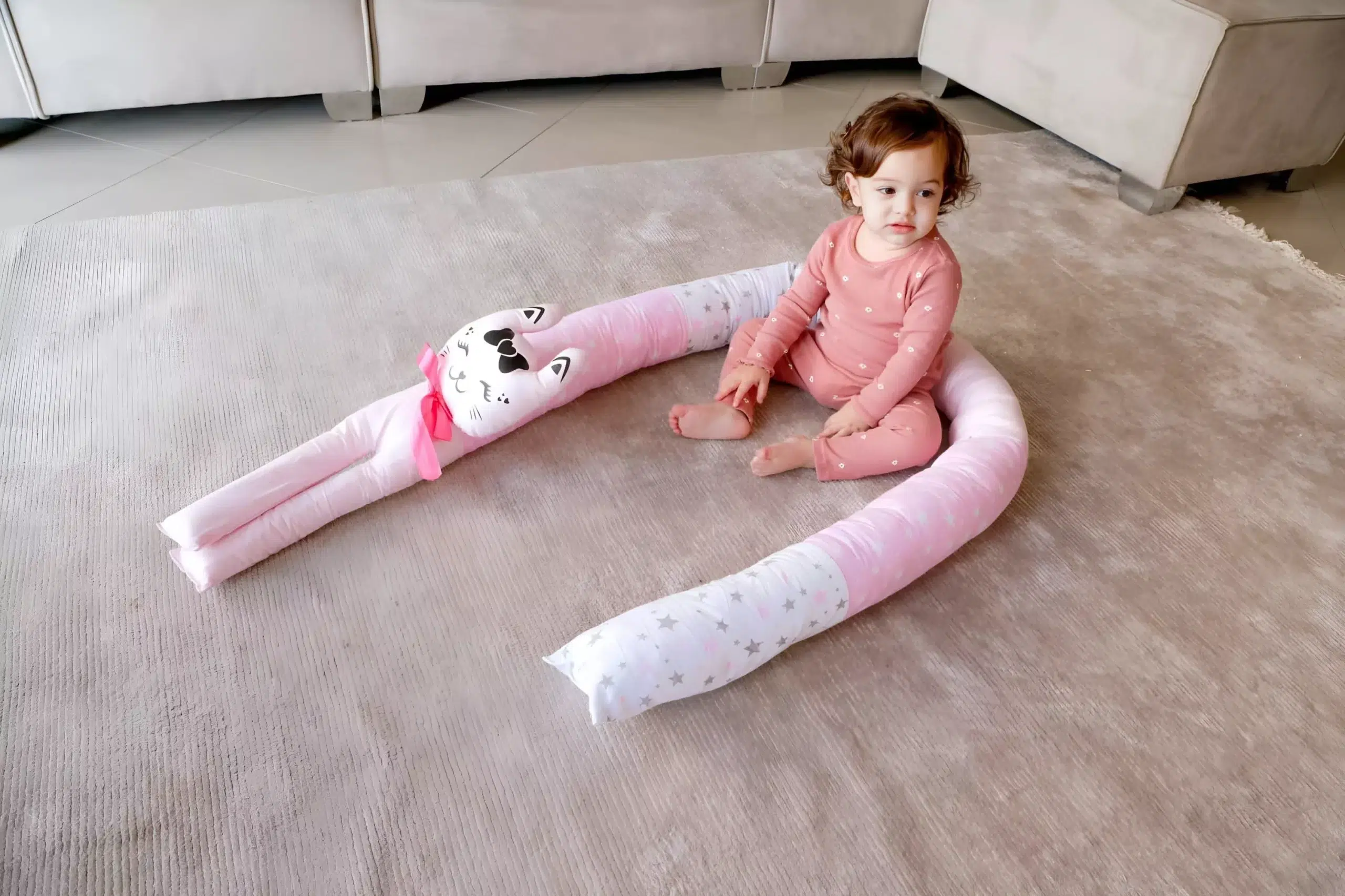 תמונה של ילדה צעירה משחקת ליד כרית בצורת חיית מחמד, עם שמלה ורודה ורקע של ספה בהירה ושטיח.